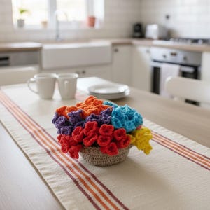 Puede incluir: Un arreglo floral de ganchillo en una pequeña cesta tejida se encuentra sobre un camino de mesa color crema con rayas naranjas y granates. Las flores son de varios colores, incluyendo rojo, naranja, azul, morado y amarillo. Dos tazas y platos blancos están en el fondo.
