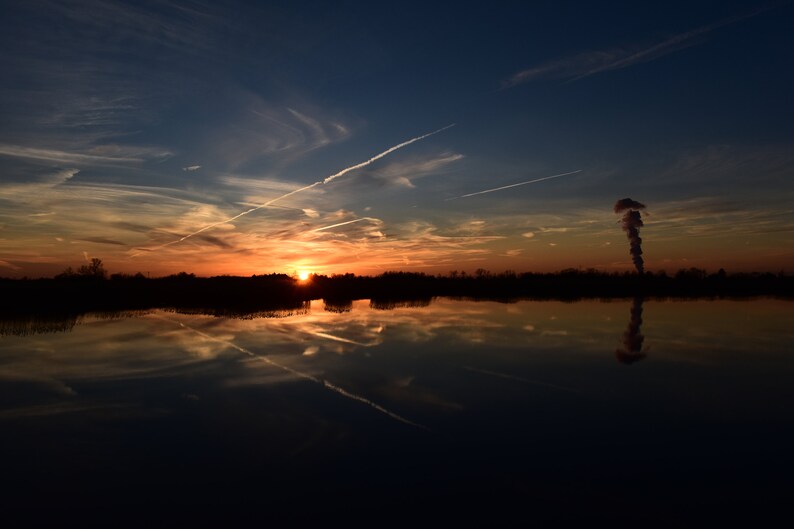 May include: A vibrant sunset over a calm body of water, reflecting the sky's colors. The sky transitions from deep blue to orange and yellow, with contrails and a plume rising from the horizon. The scene evokes tranquility.
