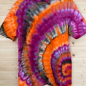 May include: A short-sleeve t-shirt featuring a vibrant tie-dye pattern in shades of bright orange, deep magenta, and dark charcoal gray. The design radiates from a spiral center point, creating a swirling, multi-colored effect across the fabric.