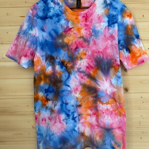 May include: A short-sleeved t-shirt with a vibrant tie-dye pattern. The shirt features a blend of colors, including blue, pink, orange, and brown, creating a unique and colorful design. The shirt is hanging on a wooden hanger.