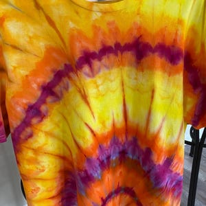 May include: A short-sleeve t-shirt featuring a vibrant tie-dye pattern in shades of bright yellow, orange, and magenta. The design radiates from the center in a sunburst style, covering the entire front of the garment with blended colors.