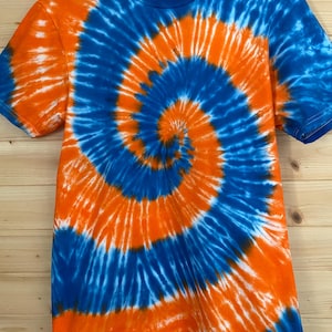 May include: A short-sleeve crew neck t-shirt featuring a vibrant tie-dye spiral pattern in bright orange and deep blue. The fabric displays a swirling design that radiates from the center, creating a bold contrast between the two colors.