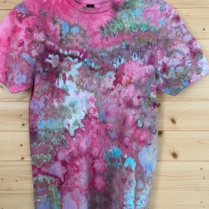 May include: A short-sleeved t-shirt with a vibrant tie-dye pattern in shades of pink, purple, blue, and green. The shirt is hanging on a wooden hanger against a light brown wooden background. The t-shirt has a crew neck and a pocket.