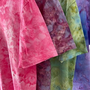 May include: A collection of four short-sleeved t-shirts featuring a tie-dye pattern. The shirts are displayed in a row, showcasing vibrant colors including bright pink, deep purple, olive green, and a blend of blue and purple hues throughout.