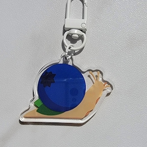 May include: A clear acrylic keychain featuring a cartoon snail with a large blue blueberry shell. The snail has a tan body, green leaves, and a white clasp. The keychain is on a white background.