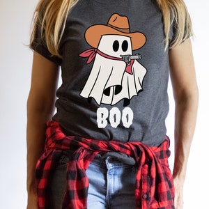 Może przedstawiać: Szary t-shirt z białym duchem w brązowym kowbojskim kapeluszu trzymającym pistolet. Duch ma czerwoną chustkę na szyi. Pod duchem nadrukowany jest napis "BOO".
