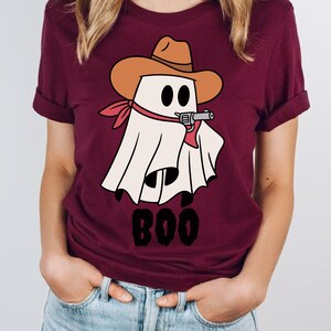 Może przedstawiać: Bordowy t-shirt z kresk&oacute;wkowym duchem w kowbojskim kapeluszu trzymającym pistolet. Duch jest biały z czarnymi oczami i czerwoną bandaną. Pod duchem nadrukowany jest napis "BOO".