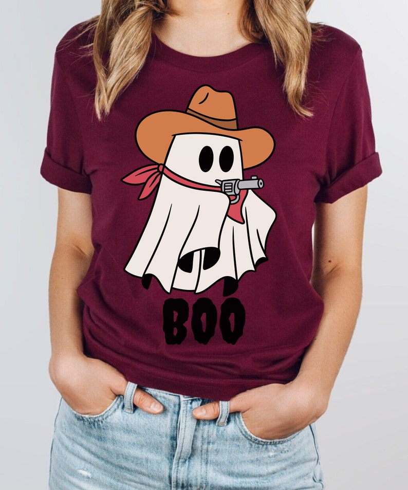 Może przedstawiać: Bordowy t-shirt z kresk&oacute;wkowym duchem w kowbojskim kapeluszu trzymającym pistolet. Duch jest biały z czarnymi oczami i czerwoną bandaną. Pod duchem nadrukowany jest napis "BOO".