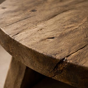 May include: Close-up of a round, rustic wooden table. The table has a natural, weathered appearance with visible wood grain and cracks. The colour is a warm, medium brown. The table's edge is thick and rounded.