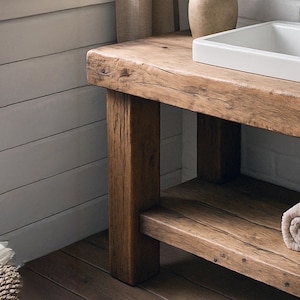 May include: A rustic wooden bathroom vanity with a natural finish. The vanity features a thick, solid wood top with a built-in white sink. The design includes a lower shelf and sturdy, square legs, showcasing the wood's grain and texture. A rolled towel rests on the shelf.
