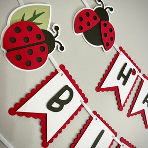 May include: A red and black ladybug birthday banner with white scalloped edges and black letters spelling "Happy Birthday".