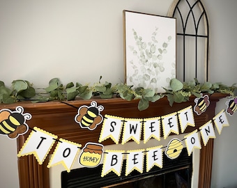 May include: A yellow and white banner with the words "Sweet To Bee One" decorated with honey pots, bees, and honeycomb. The banner is strung with black string and is hanging on a wooden mantel.