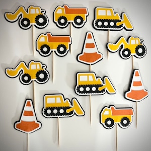 May include: A collection of yellow and orange construction-themed cupcake toppers. The toppers include images of dump trucks, excavators, and traffic cones, all outlined in black. Each topper is attached to a wooden stick.