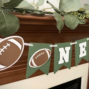 May include: Green and brown football-themed banner with the word "ONE" in white letters. The banner is decorated with brown footballs.