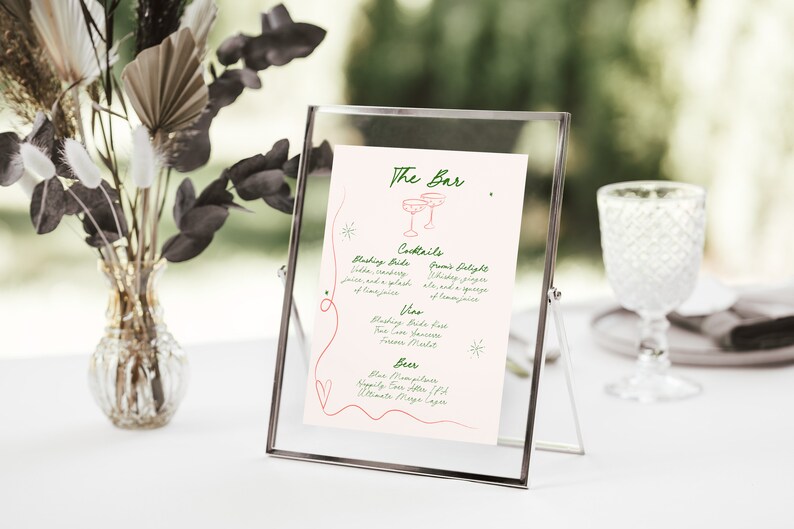 Pu&ograve; includere: Un cartello rosa e bianco con testo verde in una cornice argentata. Il cartello recita "The Bar" e elenca le opzioni di cocktail, vino e birra. Il cartello &egrave; su un tavolo bianco con un vaso di fiori secchi.