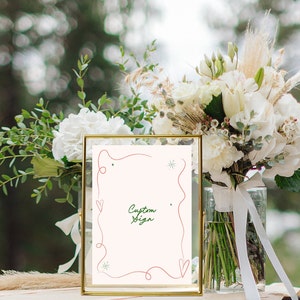 Pu&ograve; includere: Un cartello con cornice dorata, sfondo bianco e un design scritto a mano in rosso e verde. Il cartello dice "Custom Sign" e si trova su un tavolo con un mazzo di fiori bianchi in un vaso.