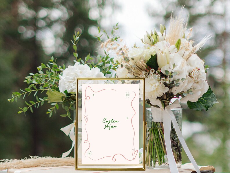 Pu&ograve; includere: Un cartello con cornice dorata, sfondo bianco e un design scritto a mano in rosso e verde. Il cartello dice "Custom Sign" e si trova su un tavolo con un mazzo di fiori bianchi in un vaso.