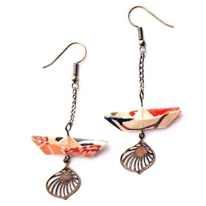 Peut inclure: Une paire de boucles d'oreilles pendantes avec des bateaux en origami en papier aux motifs rouge, blanc et noir suspendus à une chaîne avec un charme en forme de feuille de couleur bronze.