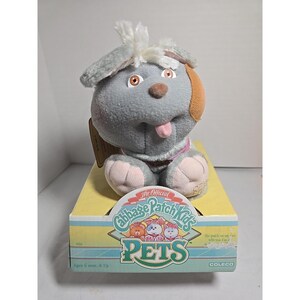 Puede incluir: Un juguete de peluche gris Cabbage Patch Kids PETS con forma de perro, con una lengua rosa y detalles marrones. El juguete está en una caja amarilla y azul con el texto "The Official Cabbage Patch Kids PETS" y "COLECO". A partir de 6 meses.