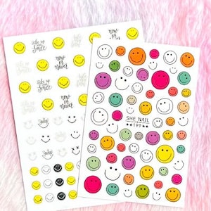 Peut inclure: Deux feuilles d'autocollants pour ongles avec des smileys colorés. La première feuille a un fond blanc avec des smileys noirs et jaunes et le texte "Wake up and smile", "Now or Never". La deuxième feuille a un fond blanc avec des smileys colorés et le texte "She Nail 199".