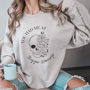May include: A light gray sweatshirt with the text "You had me at trigger warnings" printed on the front. The text is surrounded by a skull with roses.