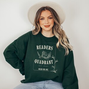 May include: A dark green sweatshirt with a white graphic design that says "Readers Quadrant Est. 2023 Read or Die". The graphic features a quill pen, an inkwell, and a stack of books.