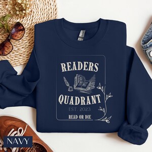 May include: Navy blue sweatshirt with a white graphic design that says "Readers Quadrant Est. 2023 Read or Die". The graphic features a quill pen, an inkwell, and a stack of books.