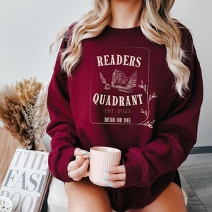 May include: A maroon sweatshirt with a white graphic design that says "Readers Quadrant Est. 2023 Read or Die". The graphic features a stack of books, a quill pen, and a feather.