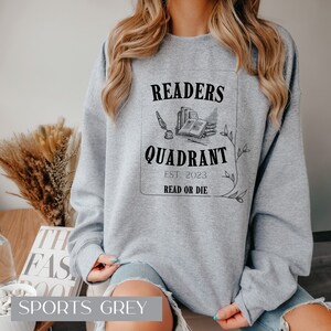 May include: A grey sweatshirt with a black graphic design that says "Readers Quadrant Est. 2023 Read or Die". The graphic features a quill pen, an open book, and a stack of books.