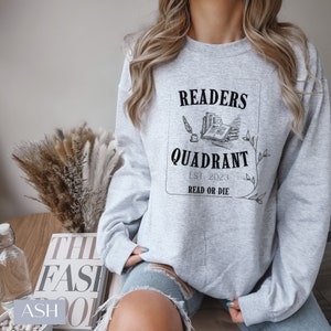 May include: A grey sweatshirt with a black graphic design that says "Readers Quadrant Est. 2023 Read or Die". The graphic includes a drawing of a quill pen and a stack of books.