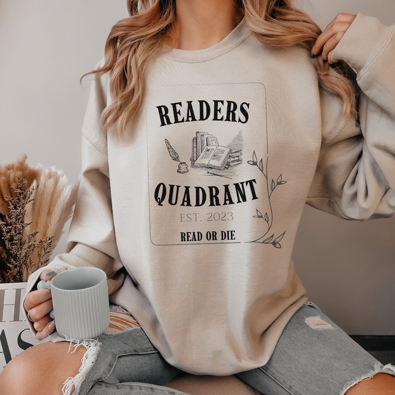 May include: A beige sweatshirt with a black graphic that says "Readers Quadrant Est. 2023 Read or Die". The graphic features a quill pen, an inkwell, and a stack of books.
