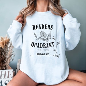 May include: A light blue sweatshirt with a graphic design that says "Readers Quadrant Est. 2023 Read or Die". The graphic features a stack of books, a quill pen, and a decorative flourish.