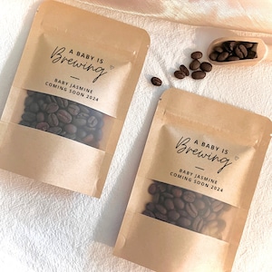 Può includere: Due sacchetti di carta marrone con finestre trasparenti riempiti di chicchi di caffè. I sacchetti hanno etichette che dicono "A Baby is Brewing" con il nome "Baby Jasmine" e l'anno "2024".