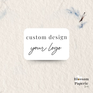 Puede incluir: Pegatina rectangular blanca con texto negro que dice "custom design your logo". La pegatina está sobre un fondo beige claro con manchas de acuarela azules y marrones. El texto "Blossom Paperie Shop" está en la esquina inferior derecha.