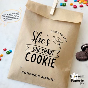 May include: Brown paper bag with a graduation theme. The bag features a black and white graphic that says "She's One Smart Cookie" with a graduation cap and the text "Class of 2024". The bag also has the text "Congrats Alison!" and a small logo that says "Blossom Paperie Shop".