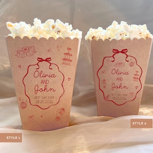 May include: Two popcorn containers filled with popcorn. The containers are light brown with a scalloped top edge. Each container has a red decorative design with the names "Olivia and John" and the date "08.25.2026".