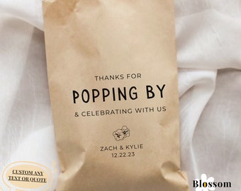 May include: A brown paper bag with the text "THANKS FOR POPPING BY & CELEBRATING WITH US" and a small illustration of popcorn. Below the text is "ZACH & KYLIE 12.22.23". The bag is on a white surface.