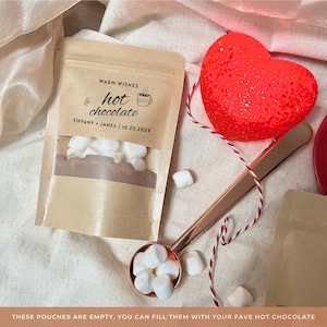 Puede incluir: Una bolsa de papel marrón con una etiqueta blanca que dice "Warm Wishes hot chocolate Tiffany + James | 12.25.2024". La bolsa está llena de mini malvaviscos y un corazón rojo está en el fondo.