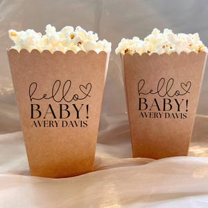 Peut inclure: Deux boîtes à popcorn en papier brun remplies de pop-corn. Chaque boîte a un dessus festonné et les mots "hello BABY!" et "AVERY DAVIS" imprimés en noir. Les boîtes sont sur une surface de couleur claire.