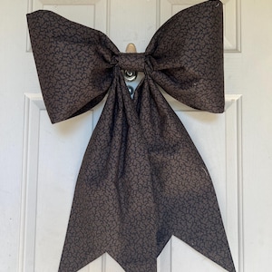 Puede incluir: Un lazo decorativo grande hecho de tela marrón con un sutil estampado floral. El lazo está sujeto a una puerta blanca. La tela está doblada y moldeada para crear un diseño de lazo clásico. Las colas del lazo tienen un borde decorativo puntiagudo.