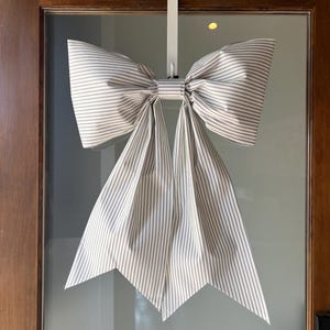 Peut inclure: Un grand nœud décoratif en tissu rayé, dans les tons de blanc et de gris, est suspendu à un crochet de porte blanc. Le nœud est centré sur une porte vitrée, le tissu tombant vers le bas dans un motif symétrique. Les rayures du nœud sont verticales.