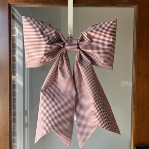 May include: A large, decorative bow made of red and white checkered fabric, hanging on a door. The bow is attached to a white door hanger. The bow's tails have a pointed, angled cut. The door is made of wood and glass.