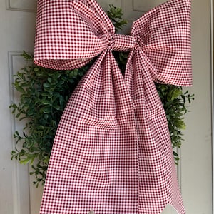 Puede incluir: Una corona decorativa con un gran lazo de cuadros rojos y blancos. El lazo es el punto focal, sobre un fondo de follaje verde. La corona está probablemente diseñada para la decoración de temporada o festiva.