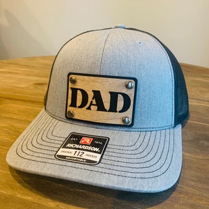 May include: A gray and black trucker hat with a wooden patch that says "DAD" on it. The hat is adjustable and has a Richardson 112 logo on the side.