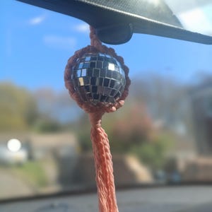 May include: A pink and silver disco ball car charm with a long tassel hanging from the rearview mirror of a car.
