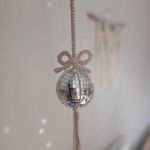 May include: A beige macrame hanging with a silver disco ball and a tassel. The disco ball is made of small mirrored tiles.