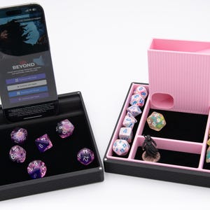 May include: A black dice tray with a phone displaying "BEYOND" and a pink dice tower. The tray holds various polyhedral dice in shades of purple and clear. The dice tower has a slot for rolling dice. A small figurine is also present.