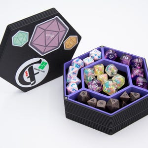 May include: A black, multi-sided dice storage box with a purple interior, filled with various colorful dice. The lid, adorned with stickers, is open beside the box. The stickers include geometric shapes and the text "crop" and "Rambou Lab".