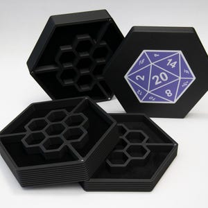 Könnte beinhalten: Schwarze sechseckige Box mit einem Wabenmuster im Inneren. Der Deckel der Box hat einen violetten 20-seitigen Würfel mit den Zahlen 2, 12, 10, 8, 14, 16, 18, 20, 1, 19, 17, 15, 6, 4, 3, 5, 9, 7, 11 und 2.