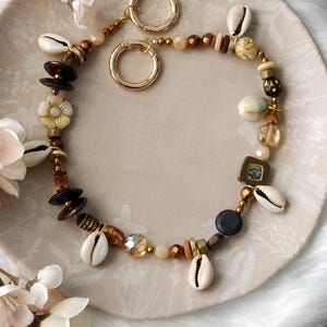 May include: A handcrafted necklace featuring a variety of beads, including seashells, wooden beads, and gold-colored metal accents. The necklace is arranged on a beige plate and is surrounded by white fur and pink flowers.